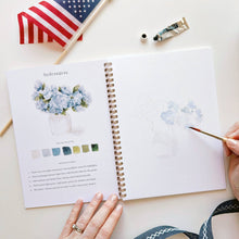 Load image into Gallery viewer, Watercolor workbook open to hydrangeas tutorial page with paint supplies and American flag