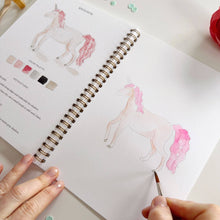 Load image into Gallery viewer, Open spiral-bound watercolor workbook showing unicorn painting tutorials with color palette and hands holding brush