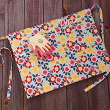Load image into Gallery viewer, Floral patterned half apron with tool pocket containing pink gardening gloves on wooden surface