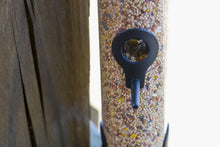 Load image into Gallery viewer, Matte black bird seed feeder filled with mixed seeds hanging outdoors near wooden fence