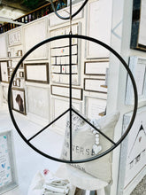 Load image into Gallery viewer, Black metal peace sign wall decor hanging in rustic white display room with gallery frames