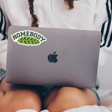 Load image into Gallery viewer, Green leaf-shaped 'Homebody' sticker on MacBook laptop held by person in bed