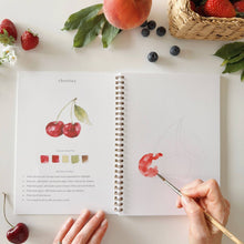 Load image into Gallery viewer, Open watercolor workbook showing cherry painting tutorial with hands holding brush and fresh fruit