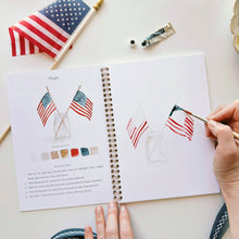 Load image into Gallery viewer, Hands painting American flags in watercolor workbook with art supplies and reference flag nearby