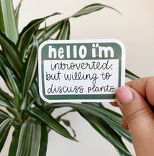 Load image into Gallery viewer, Hand holding green and white sticker reading 'hello i'm introverted, but willing to discuss plants' near houseplant