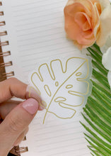 Load image into Gallery viewer, Hand holding clear vinyl sticker with golden monstera leaf outline design