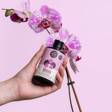 Load image into Gallery viewer, Hand holding Bloom fertilizer bottle with purple spotted orchid flowers on pink background