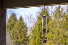 Load image into Gallery viewer, Copper finish tube bird feeder hanging from covered porch with green trees background