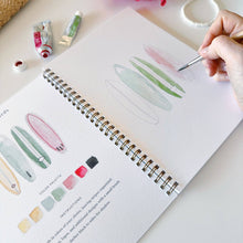 Load image into Gallery viewer, Hand painting tropical leaves in spiral-bound watercolor workbook with paint tubes and brush nearby