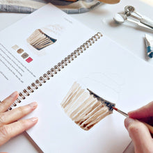 Load image into Gallery viewer, Hands painting watercolor cupcake in spiral-bound baking workbook with color swatches and measuring spoons