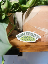 Load image into Gallery viewer, Homebody sticker with green leaf design placed on wooden surface near houseplants
