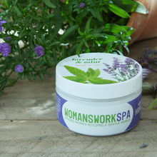 Load image into Gallery viewer, WOMANSWORKSPA lavender mint hand cream jar with purple and white labeling surrounded by flowering herbs