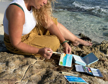 Load image into Gallery viewer, Woman painting watercolor seascape with portable travel art kit on rocky coastline
