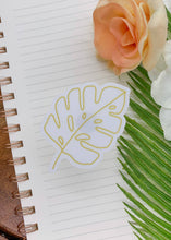 Load image into Gallery viewer, Clear vinyl sticker with golden leaf outline on lined notebook with flowers and palm frond