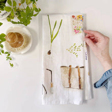 Load image into Gallery viewer, White cotton tea towel with botanical garden illustrations including plants, basket, and wildflower seed packet