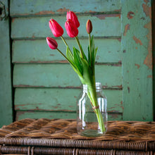 Load image into Gallery viewer, Fresh salmon-red tulips arranged in clear glass jar on wicker basket against weathered green backdrop