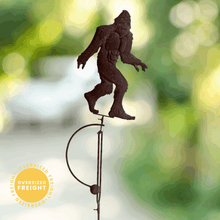 Load image into Gallery viewer, Dark metal Bigfoot silhouette garden stake with balance rocker mechanism against blurred green background