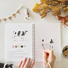 Load image into Gallery viewer, Open watercolor workbook showing cow painting tutorial with hands holding brush, surrounded by art supplies