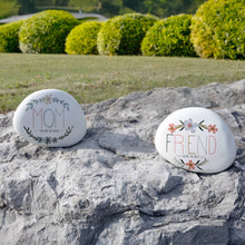 Load image into Gallery viewer, Two white decorative garden stones with floral designs reading MOM and FRIEND on rocky surface