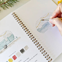 Load image into Gallery viewer, Hand painting a vintage camper trailer in a spiral-bound watercolor workbook with yellow string lights