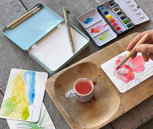 Load image into Gallery viewer, Portable watercolor painting kit with paints, brushes, and floral artwork on wooden surface