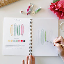 Load image into Gallery viewer, Open watercolor workbook showing surfboard painting tutorial with color palette and brush in hand