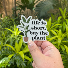 Load image into Gallery viewer, Hand holding white sticker with plant illustration reading 'life is short, buy the plant' against green foliage background