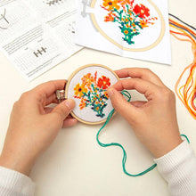 Load image into Gallery viewer, Hands working on colorful flower cross stitch embroidery in wooden hoop with pattern instructions