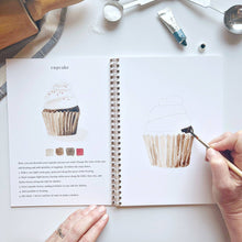 Load image into Gallery viewer, Open watercolor tutorial workbook showing cupcake painting lesson with brush and art supplies