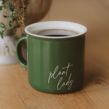 Load image into Gallery viewer, Green ceramic coffee mug with 'plant lady' text filled with coffee on wooden surface