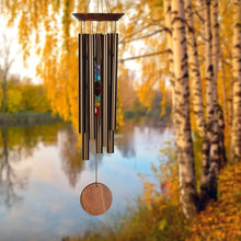 Load image into Gallery viewer, Large bronze wind chime with seven chakra-colored stones hanging in autumn forest setting