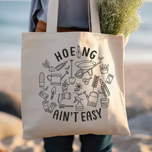 Load image into Gallery viewer, Canvas tote bag with 'Hoeing Ain't Easy' text and gardening tool illustrations