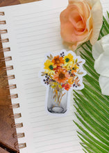 Load image into Gallery viewer, Clear vinyl sticker featuring colorful wildflowers in glass mason jar on notebook