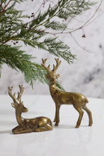 Load image into Gallery viewer, Two brass-finish deer figurines, one standing and one resting, on marble surface with evergreen branches.