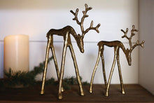 Load image into Gallery viewer, Two gold metal reindeer figurines with antlers displayed on a wooden shelf with greenery and candle
